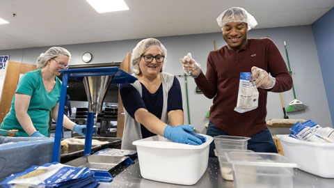 Two COD Cares volunteers wearing a head cap and gloves, sift flour in tubs. 