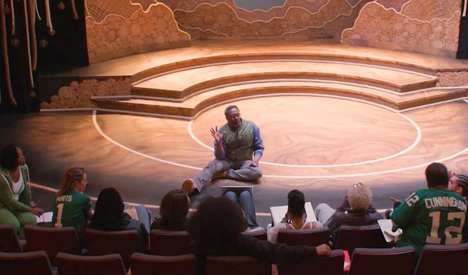 a man sitting on a small stage while students sit in the front row and look on